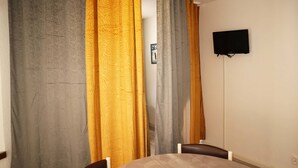Een kamer met een ronde tafel, twee stoelen, een aan de muur bevestigde televisie en gordijnen in grijs, geel en wit.
