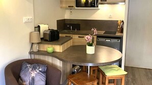 Een compacte keuken met een magnetron, broodrooster en spoelbak. Er is een klein eethoekje met een ronde tafel en twee krukken. Een stoel staat in de buurt van de keuken.