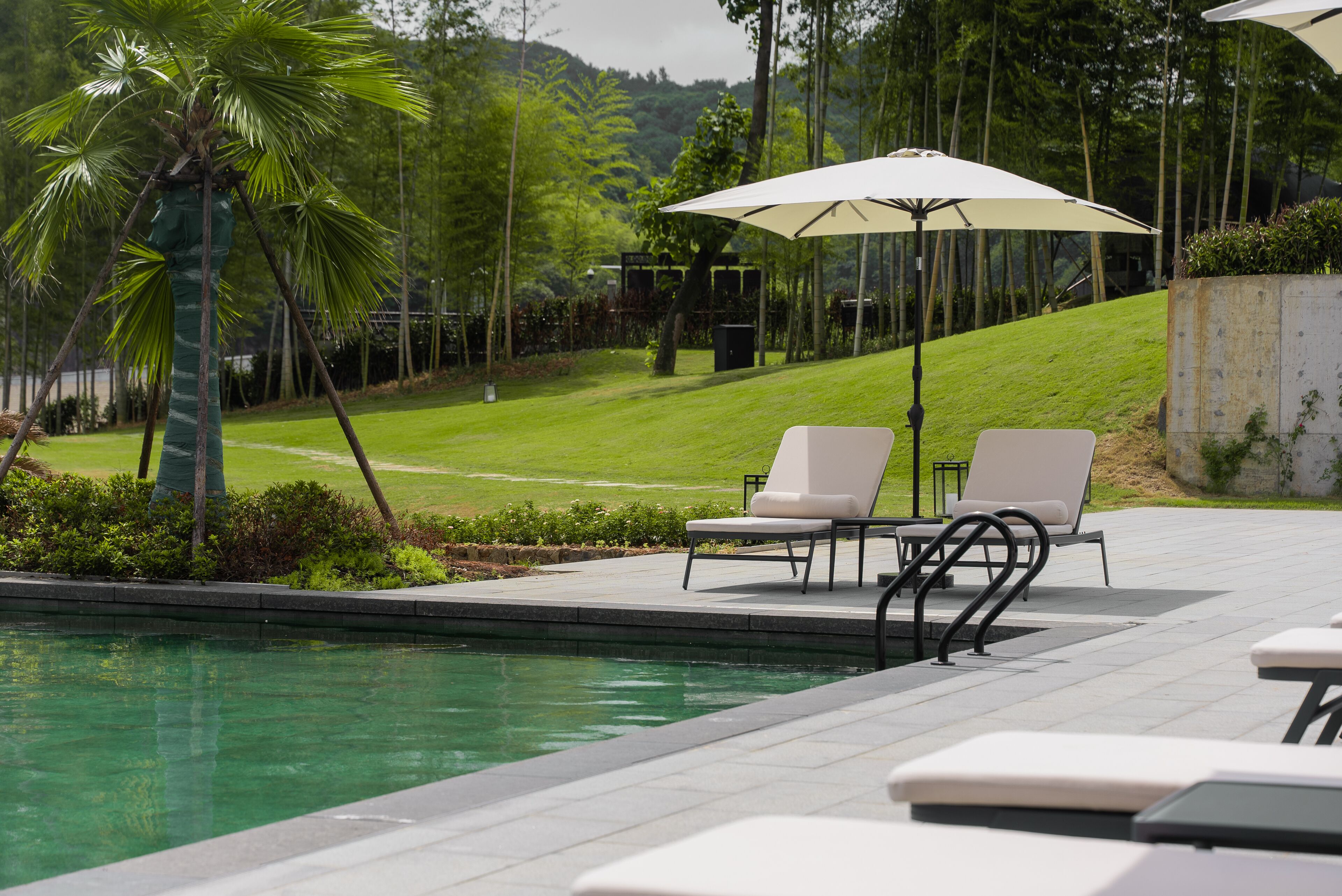 Un espace aménagé au bord de la piscine, avec des chaises longues et un grand parasol, entouré de verdure et offrant une vue sur une colline.