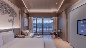 A modern hotel room with a large bed, a sofa, a dining table, and a balcony with a scenic view. - Grand Dongshan (Shaoxing)