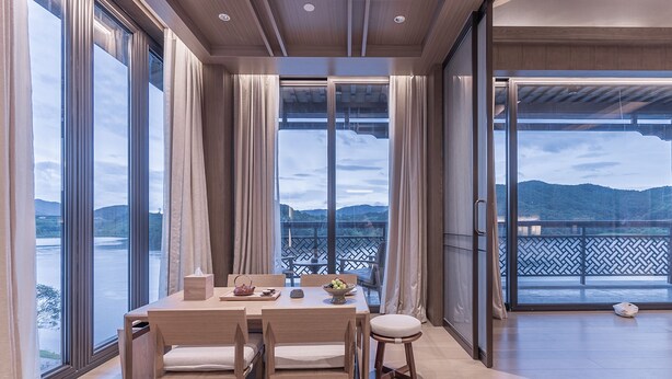 A dining area with a table set for two, large windows with curtains, and a view of a mountainous landscape. - Grand Dongshan (Shaoxing)