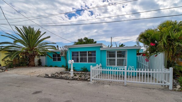 A turquoise two-story house with a white picket fence, surrounded by tropical plants and a gravel driveway.
