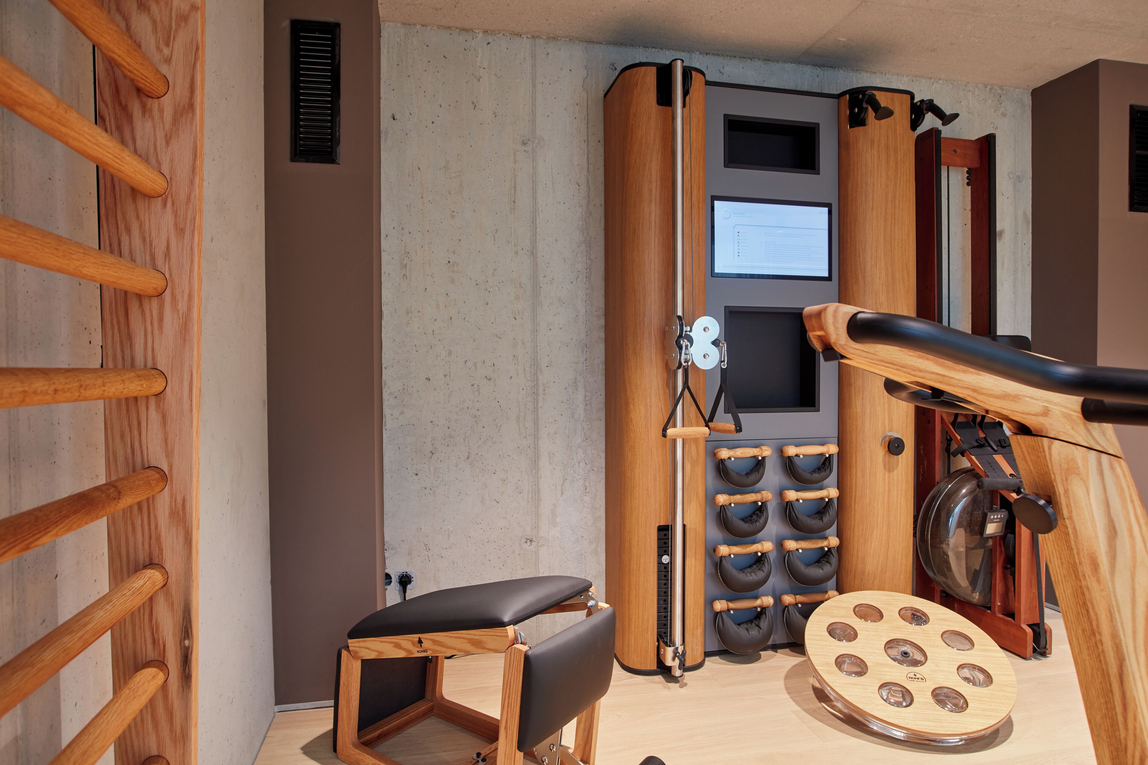 A room with a wooden bench, a wooden ladder, a wall-mounted exercise machine, and a circular wooden object.