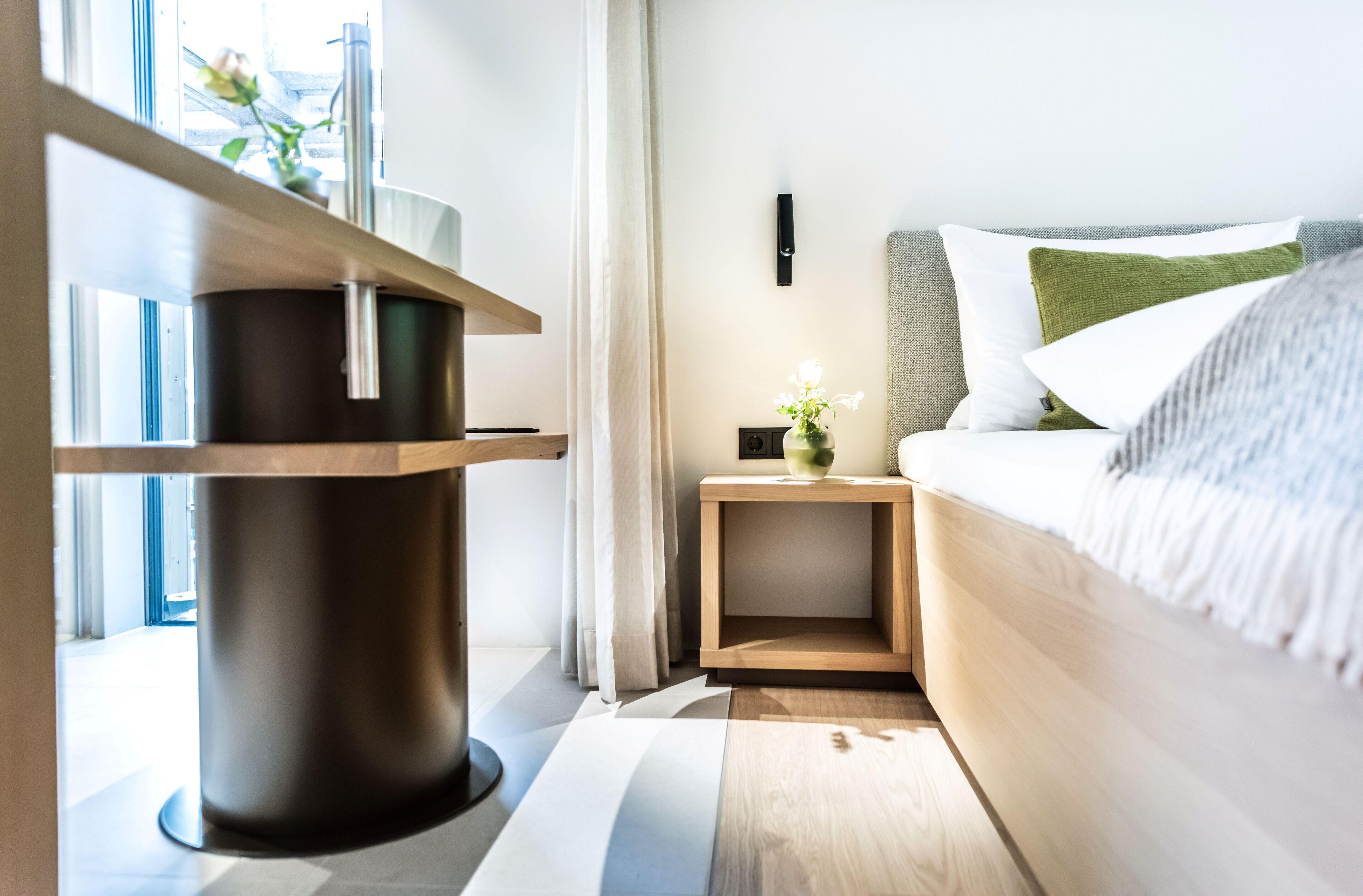 A modern bedroom with a wooden bedside table, a small nightstand with a vase of flowers, and a bed with white and green pillows.