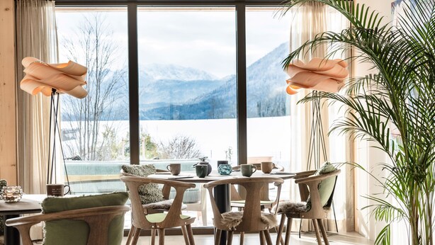 A dining area with a table and chairs, a large window with a mountain view, and a potted plant. - Hotel BERGEBLICK  (Bad Toelz)