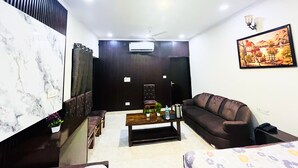 A living room with a dark brown sofa, a wooden coffee table, a marble accent wall, a ceiling fan, and a painting on the wall. - Roomshala 005 Tom & Jerry (New Delhi)