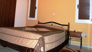 Um quarto com cama, ar condicionado, janela com persianas, mesa de cabeceira e criado-mudo.
