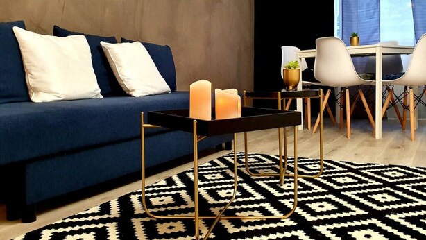 Ein modernes Wohnzimmer mit einem blauen Sofa, zwei weißen Kissen, einem schwarzen Couchtisch mit Kerzen und einem gemusterten Teppich.