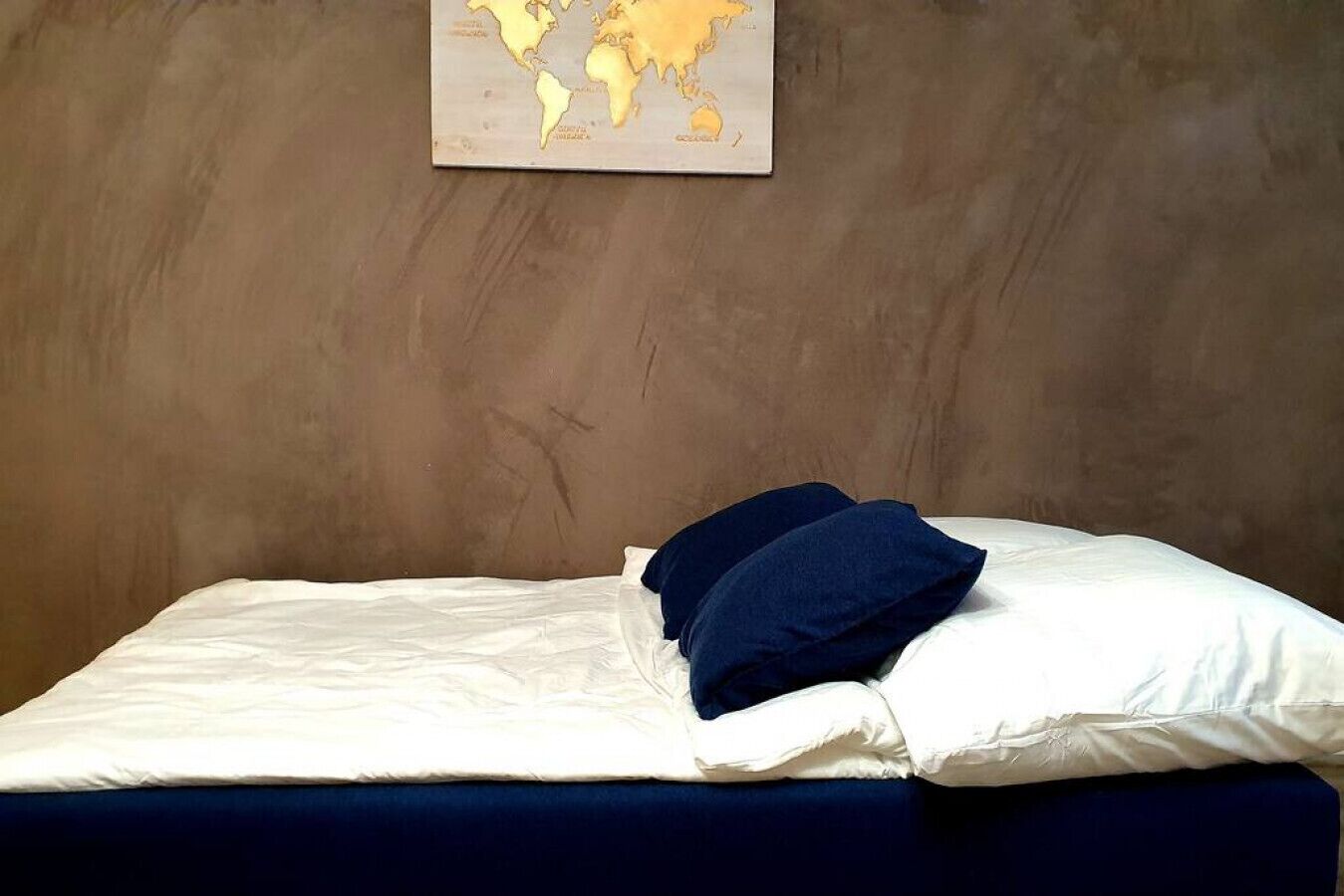 Un lit simple avec une literie blanche et un oreiller bleu foncé, placé devant un mur texturé sur lequel est accrochée une carte du monde.