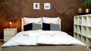 Ein ordentlich bezogenes Bett mit weißer Bettwäsche und gemusterten Kissen, eine Nachttischlampe und eine Wand mit gerahmten Bildern.
