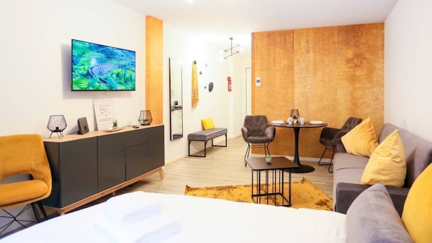 A modern hotel room with a bed, a sofa, a dining table, a TV, and a desk.
