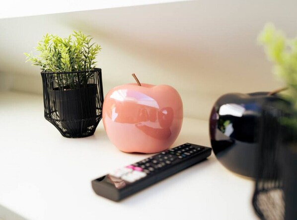 A remote control, a pink apple-shaped container, and a black vase with green plants on a white surface. - Noble Suite mit perfektem Wohn- und Arbeitskomfort (Gießen)