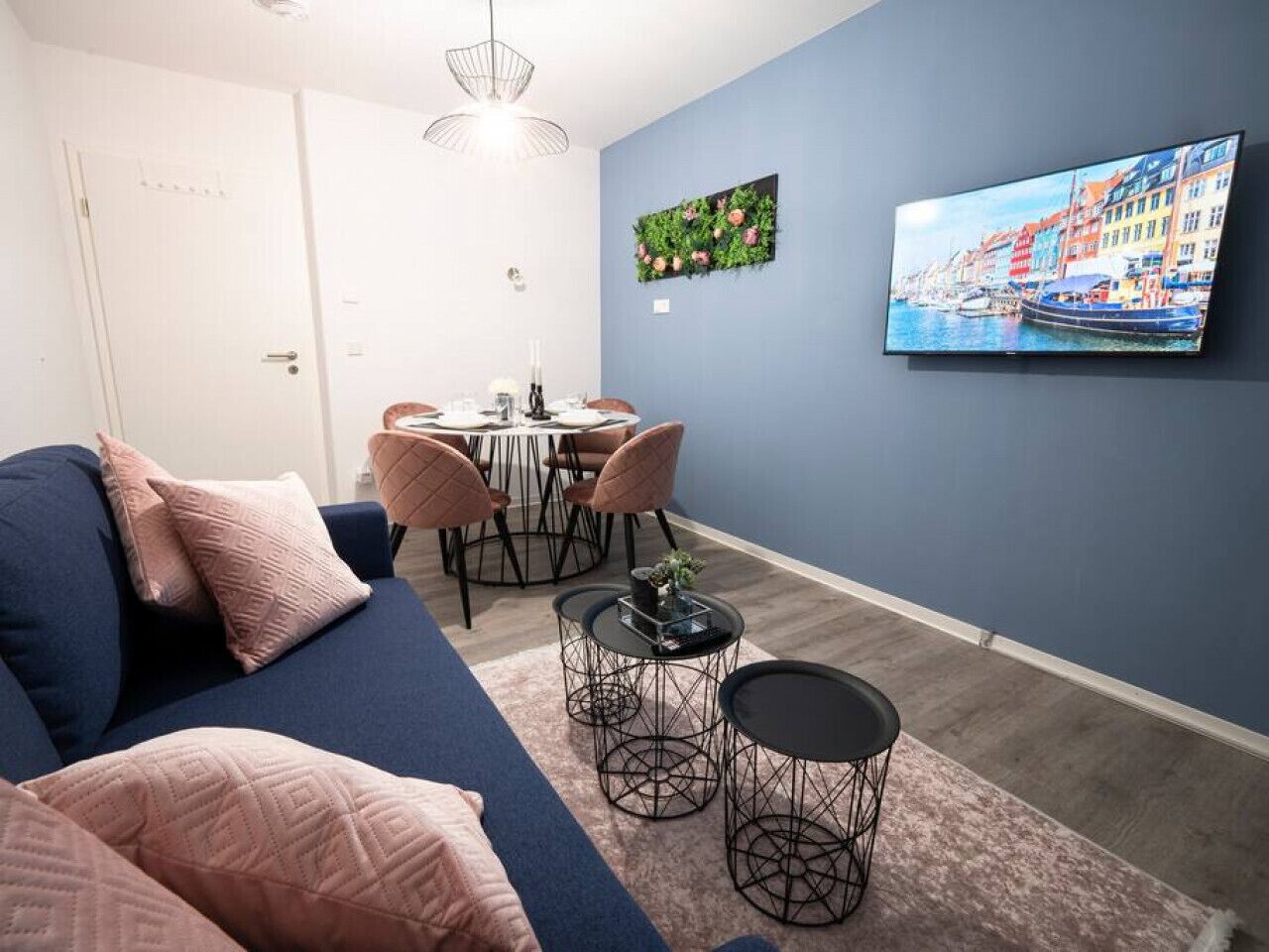 Ein modernes Wohnzimmer mit einem blauen Sofa, einem Esstisch mit Stühlen, einem an der Wand montierten Fernseher und einem Wandbild.