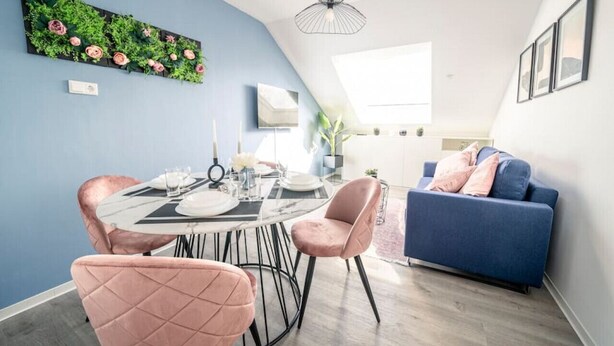 A modern dining area with a round table, pink upholstered chairs, and a blue sofa. - Noble Suite mit perfektem Wohn- und Arbeitskomfort (Gießen)