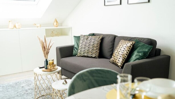 Een moderne woonkamer met een grijze bank, een rond bijzettafeltje met gouden accenten, en een eethoek met een witte tafel gedekt voor twee personen.
