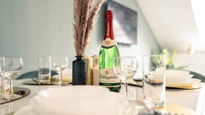 Een eettafel gedekt met witte borden, bestek en glazen, een fles Rothäppchen, en een decoratieve vaas met pluimgras.