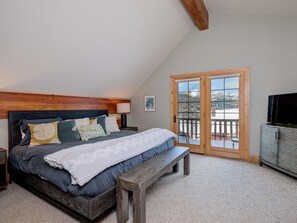 A bedroom with a large bed, wooden headboard, and a bench. There is a balcony with a view of snow-covered mountains.