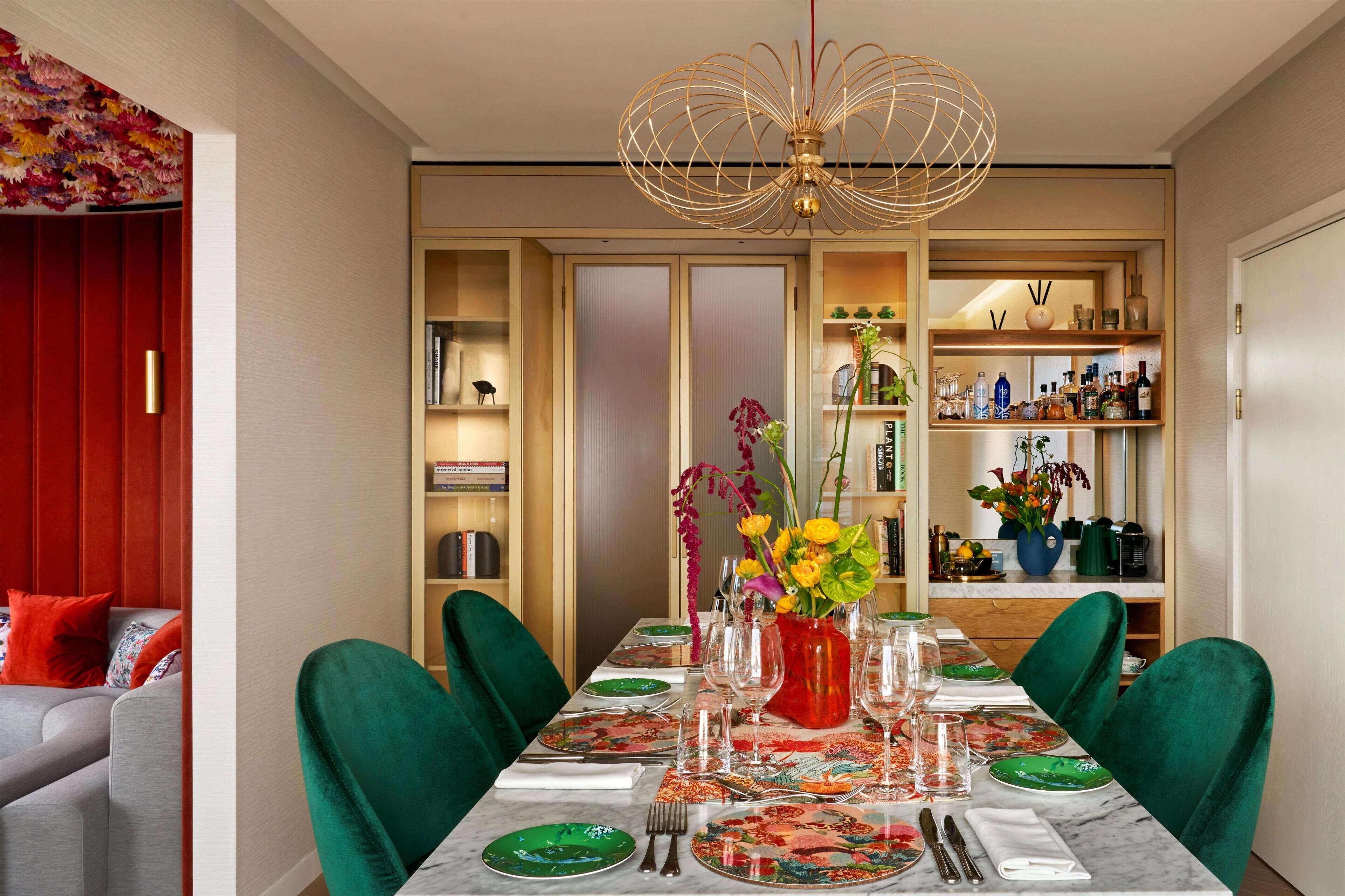 A dining area with a table set for four, green chairs, a chandelier, and a bookshelf with various items.