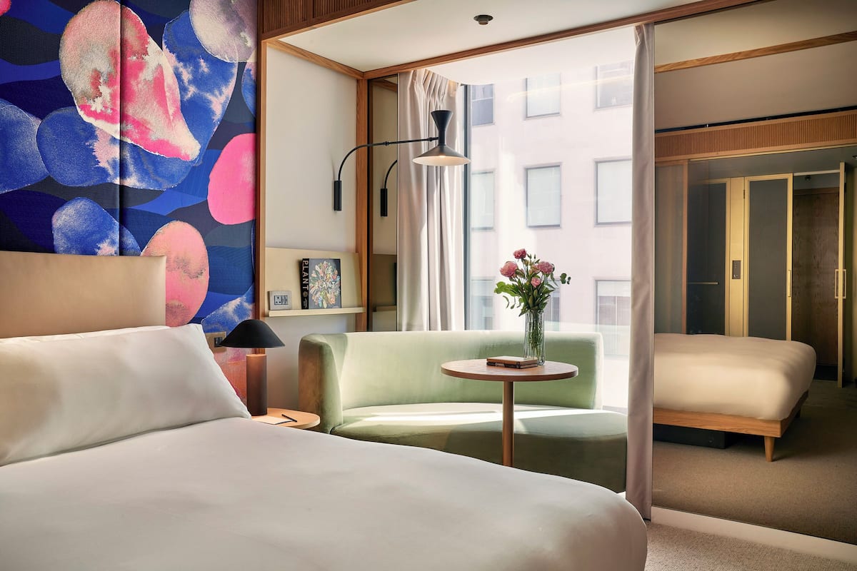 A hotel room with a bed, a green armchair, a small table with a vase, and a window with curtains.