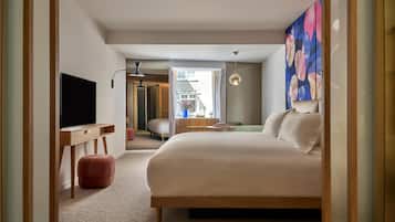 A modern hotel room with a large bed, a desk with a flat-screen TV, a small seating area, and a view of the city.