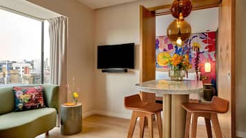 Ein modernes Wohnzimmer mit einer Couch, einem runden Tisch, zwei Stühlen, einem an der Wand befestigten Fernseher und Blick auf die Stadt durch große Fenster.