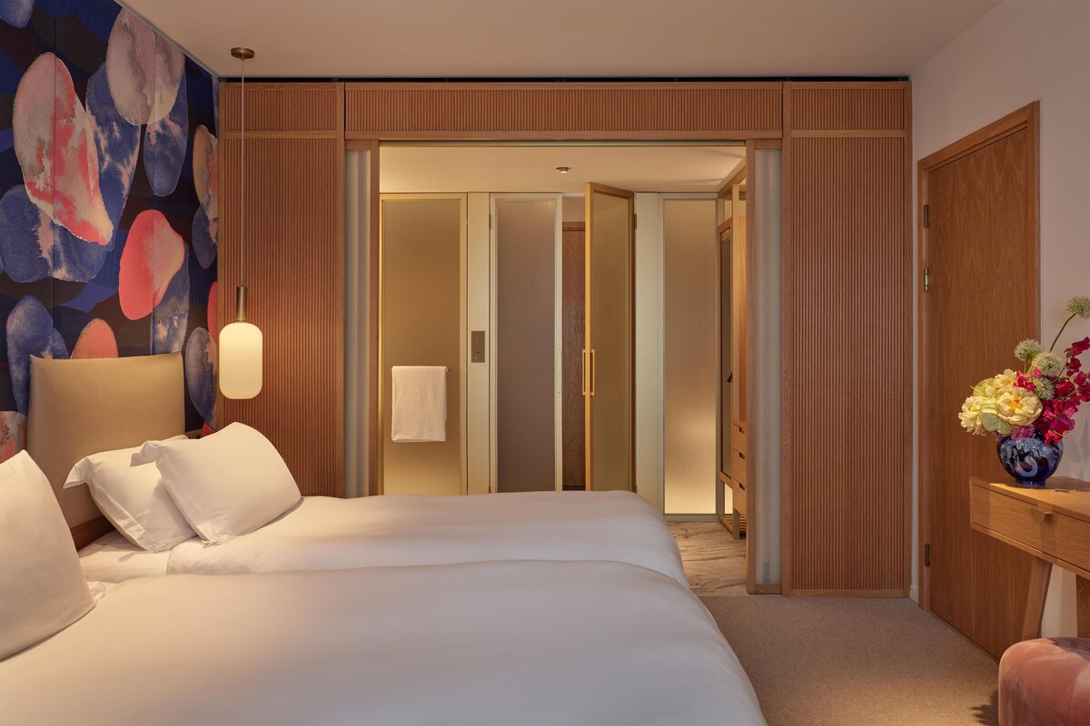 A hotel room with a large bed, a wooden wardrobe, a desk with a vase of flowers, and a wall with abstract art.