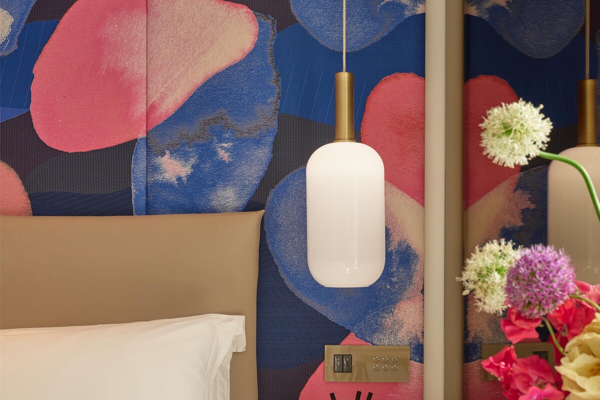 A hotel room with a bed, a wall-mounted air purifier, a small table with a decorative item, and a vase with flowers.