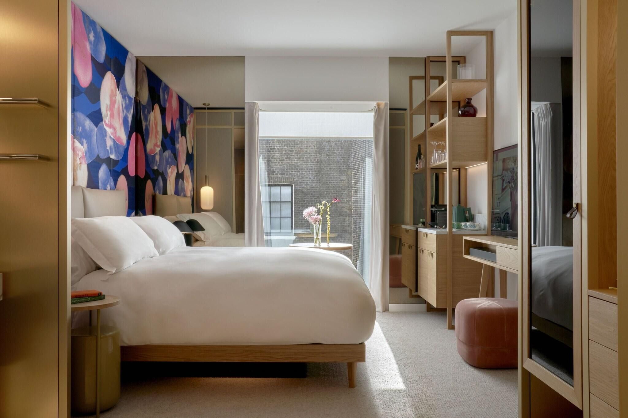Habitación de hotel moderna con una cama grande, un escritorio, un televisor y vista a una pared de ladrillo a través de una ventana.