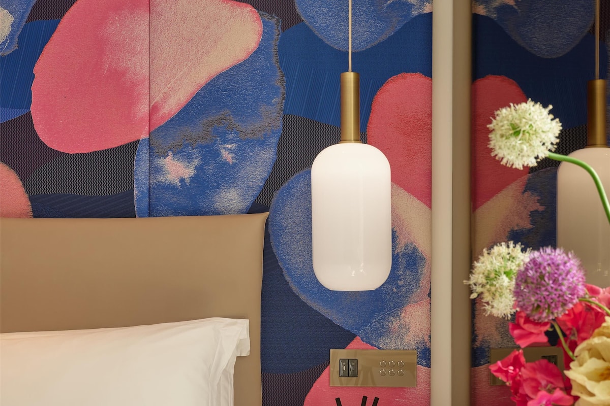 A hotel room with a bed, a wall-mounted air purifier, a small table with a decorative item, and a vase with flowers.