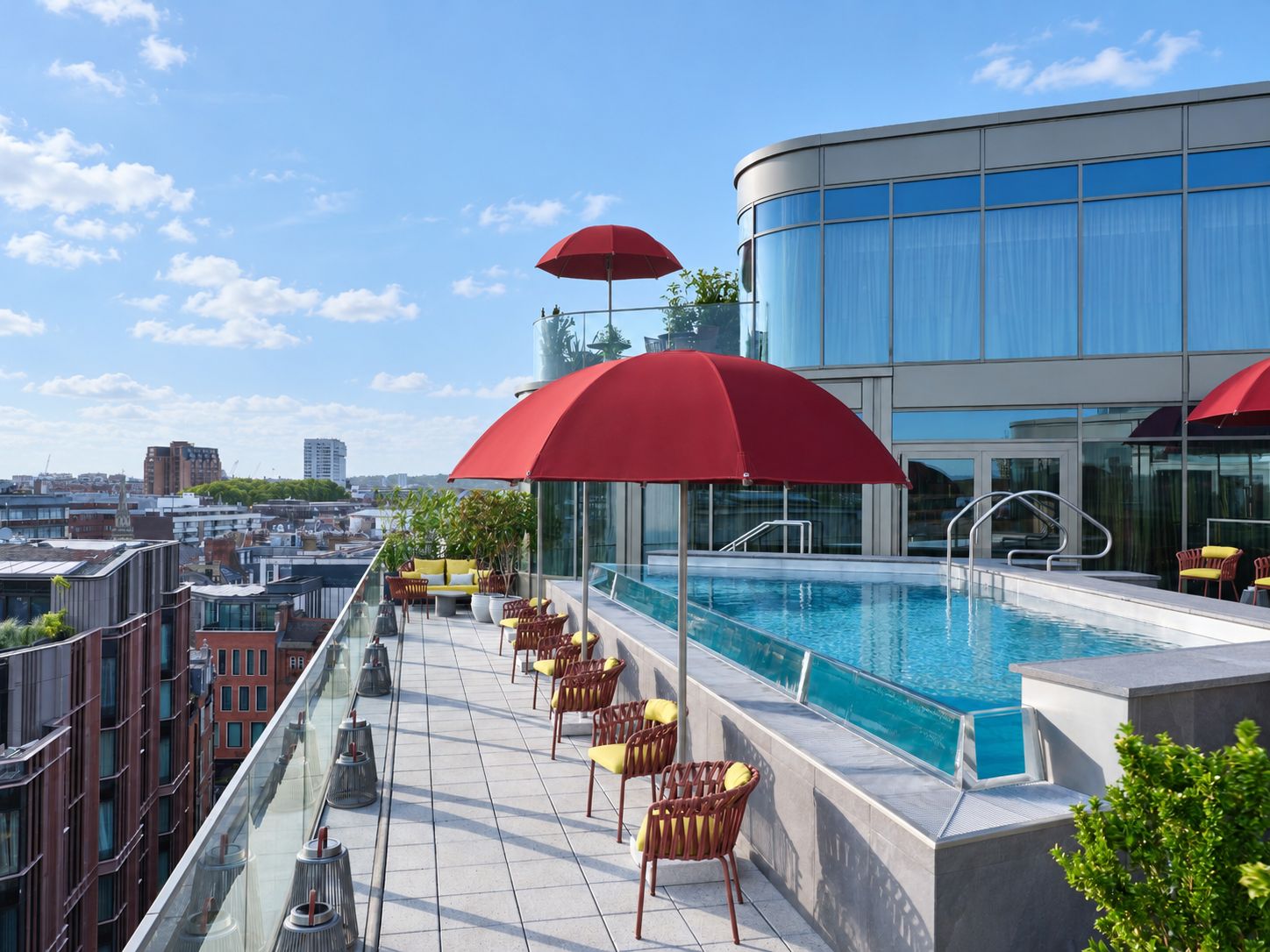A rooftop pool area with a glass balustrade, red umbrellas, and wicker chairs.