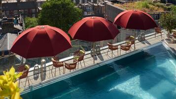 A rooftop pool with red umbrellas and wicker chairs, overlooking a cityscape.