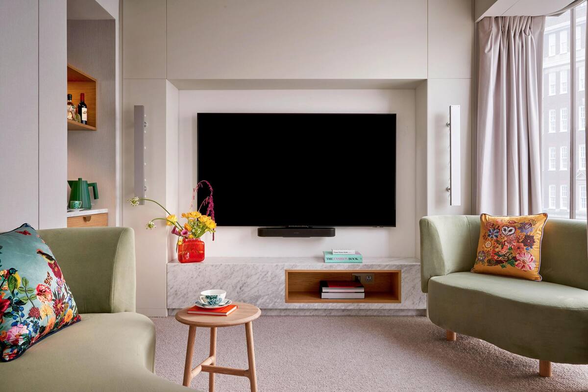 A modern living room with a flat-screen TV mounted on the wall, two sofas with colorful cushions, a small wooden side table with a cup and book, and a vase with flowers.