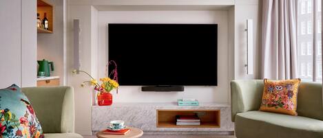 A modern living room with a flat-screen TV mounted on the wall, two sofas with colorful cushions, a small wooden side table with a cup and book, and a vase with flowers.