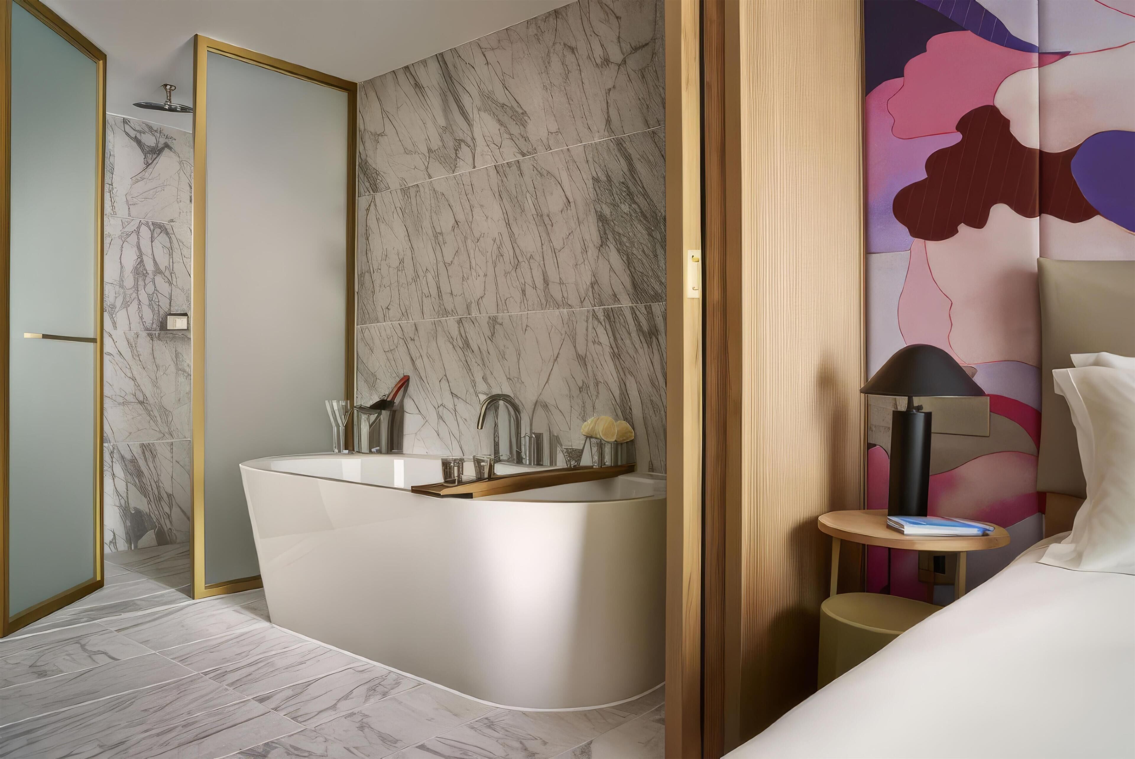 A modern bathroom with a freestanding bathtub, a mirror, and a small bedside table with a lamp.