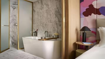 A modern bathroom with a freestanding bathtub, a mirror, and a small bedside table with a lamp.