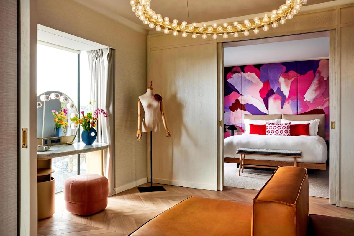 A modern hotel room with a vanity, mirror, and flowers; a bed with a colorful headboard; and a large window.