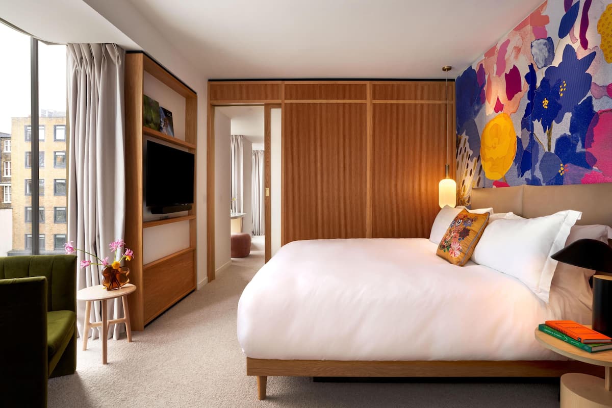 A hotel room with a large bed, a TV, a small table with a vase, and a colorful wall mural.