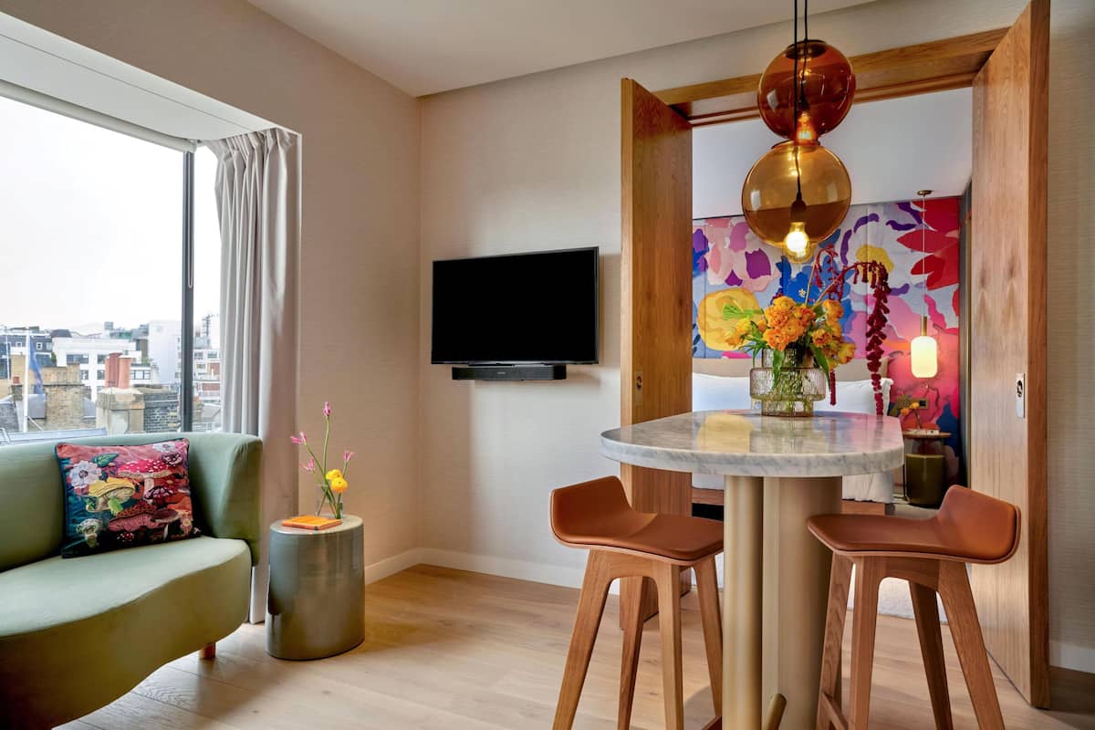 A modern living room with a sofa, a round table with two chairs, a TV mounted on the wall, and a city view through large windows.