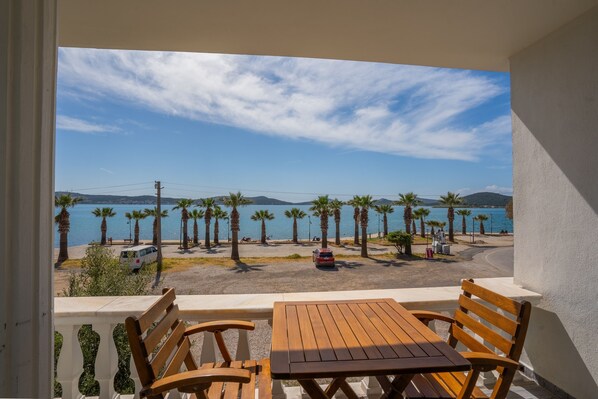 Un balcon avec une table et des chaises en bois, donnant sur une plage bordée de palmiers et offrant une vue sur la mer.