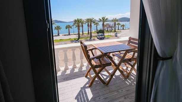 Balcón con mesa y sillas de madera, con vista a una playa con palmeras y cielo despejado.