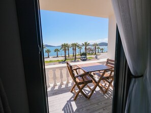 Un balcon avec une table et des chaises en bois, donnant sur une plage bordée de palmiers et offrant une vue sur un ciel dégagé.