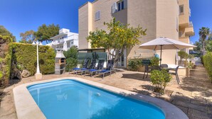 A swimming pool with lounge chairs and umbrellas, surrounded by a building with balconies and a clear blue sky. - Playamar by Rentallorca (Puerto de Alcudia)