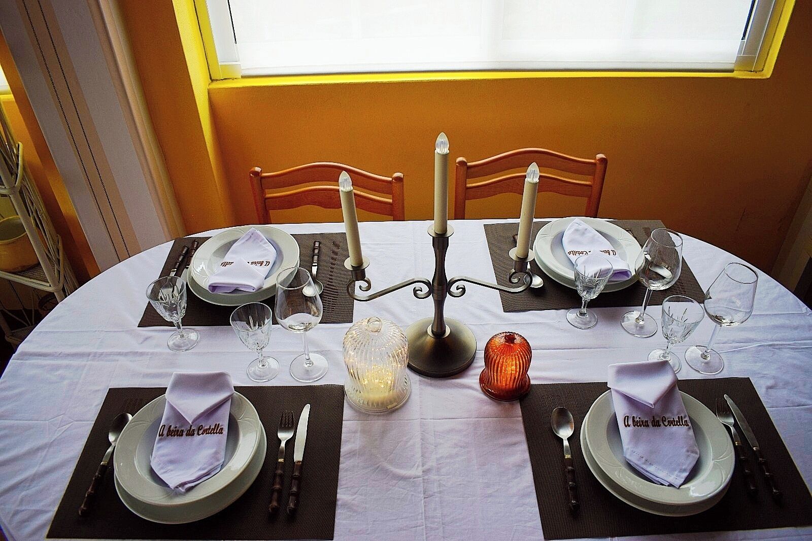 Mesa de comedor dispuesta para cuatro personas, con mantel blanco, individuales negros y cubiertos.