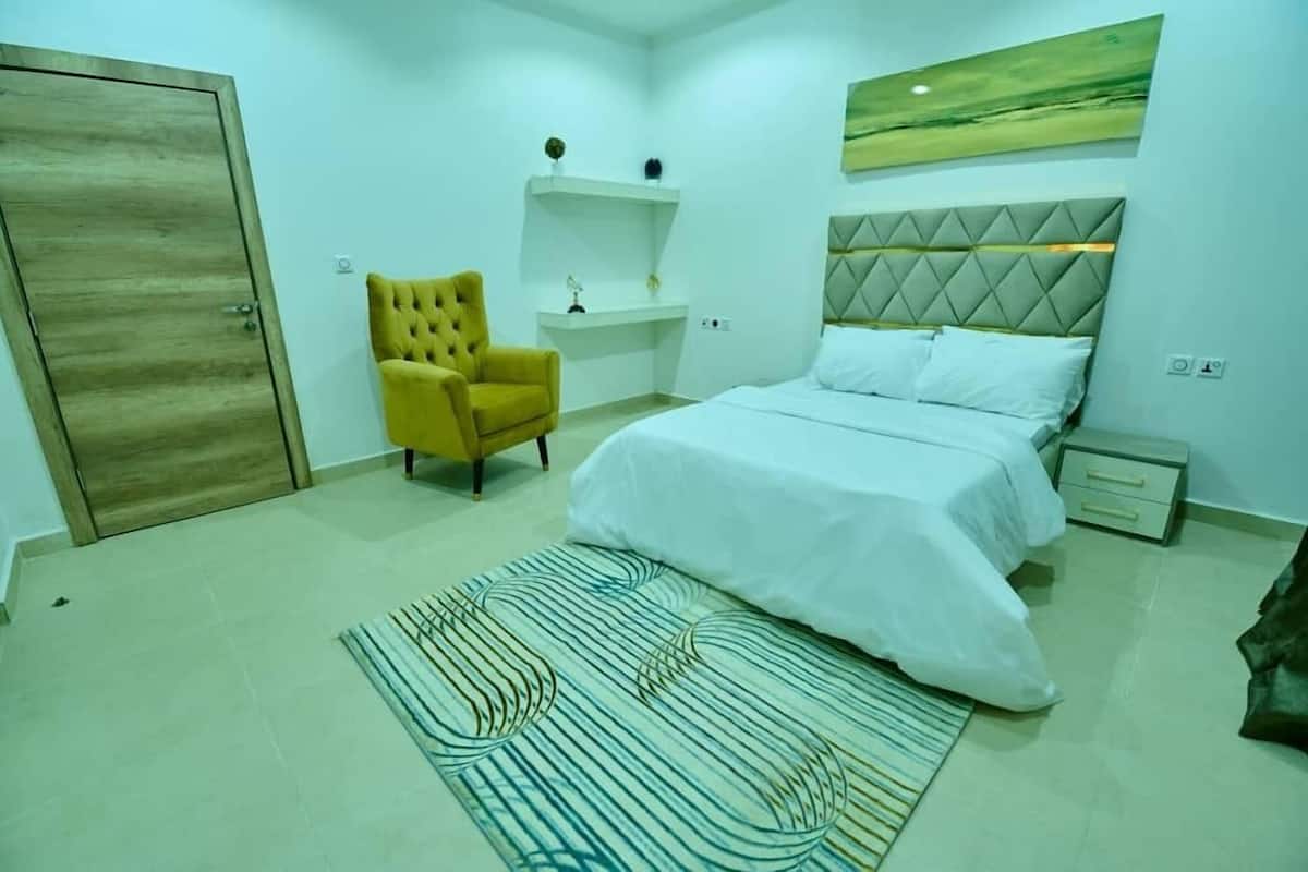 A bedroom with a bed, a yellow armchair, a bedside table, and a wall-mounted shelf.