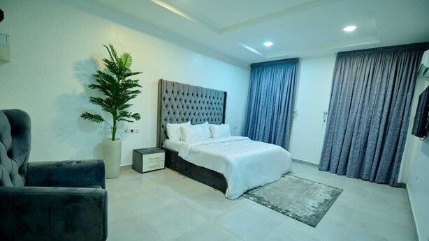 Quarto moderno com cama, poltrona, planta em vaso e uma janela grande com cortinas.