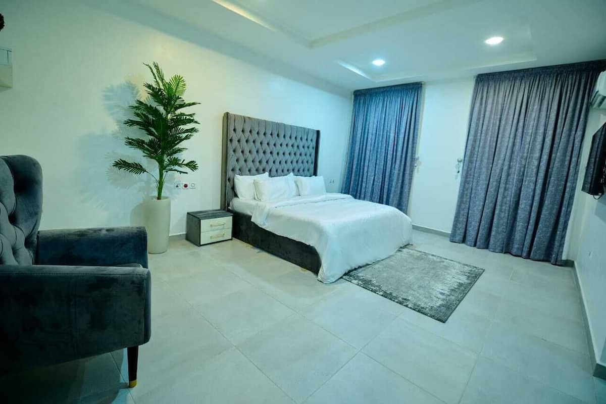 A modern bedroom with a bed, a chair, a potted plant, and a large window with curtains.