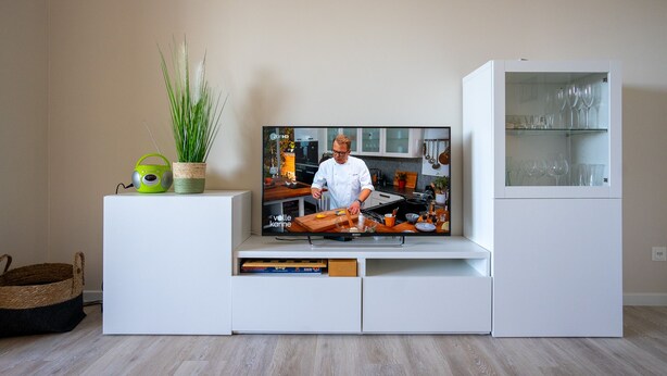 A modern living room with a flat-screen TV on a white entertainment unit, a kitchen area, and a glass-fronted cabinet with wine glasses. - Muschelbank Wohnung 59 (Büsum)