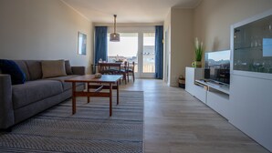 A modern living room with a sofa, a dining table, and chairs. There is a TV on a white cabinet, and a large window with curtains. - Muschelbank Wohnung 59 (Büsum)