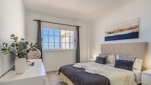 Une chambre à coucher avec un lit, une table de chevet, une commode, une fenêtre avec des rideaux, une plante en pot et un tableau accroché au mur.