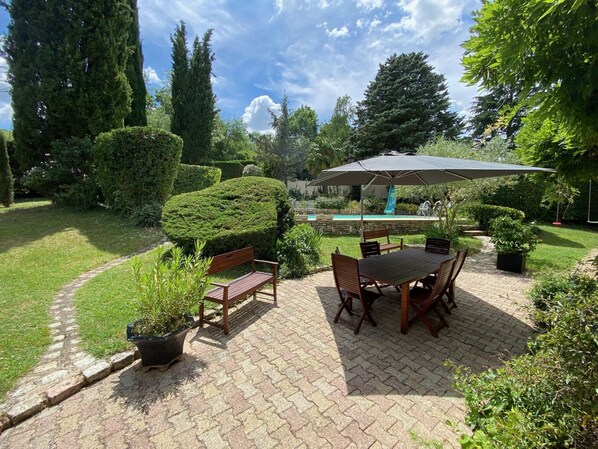 Un espace patio aménagé avec une table et des chaises, entouré de haies bien entretenues et avec une piscine en arrière-plan.